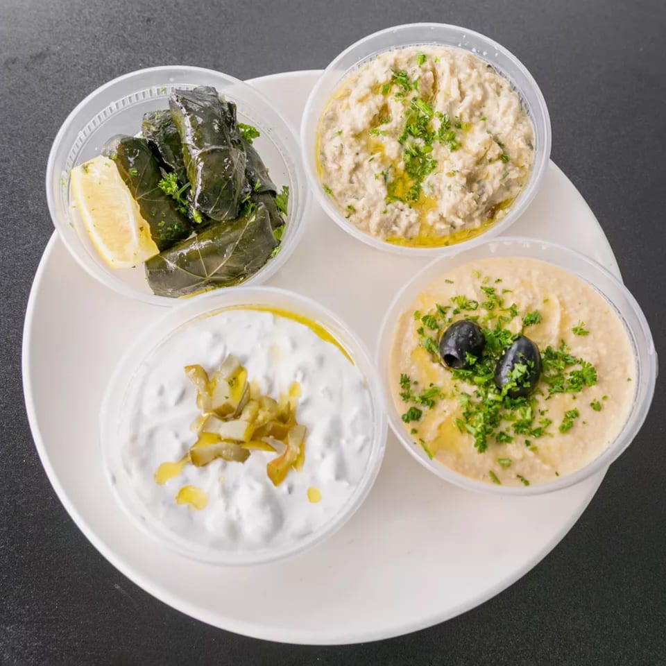 Four clear plastic containers of Mediterranean dips and dolmades on a white plate.