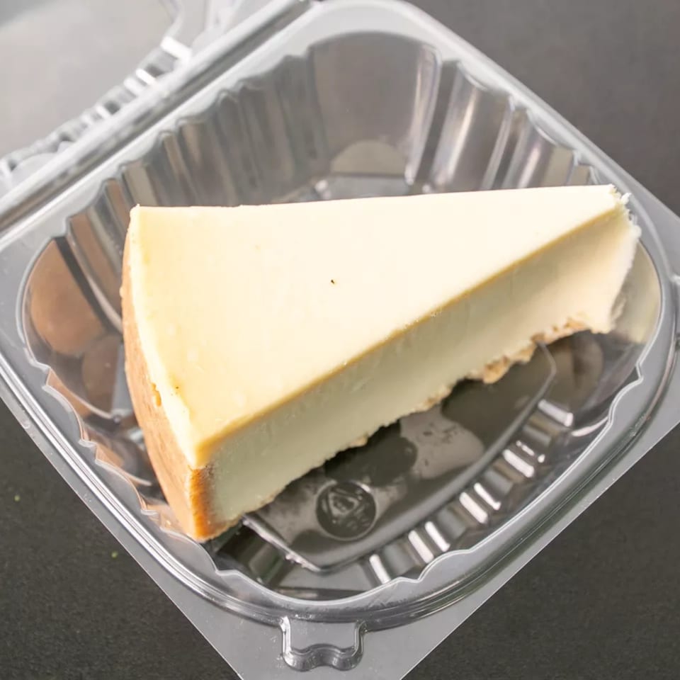 A slice of plain cheesecake with a graham cracker crust sits in a clear plastic to-go container.