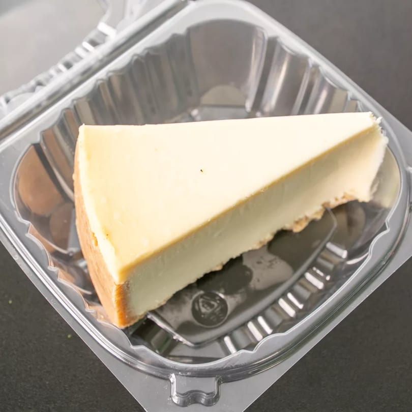 A slice of plain cheesecake with a graham cracker crust sits in a clear plastic to-go container.