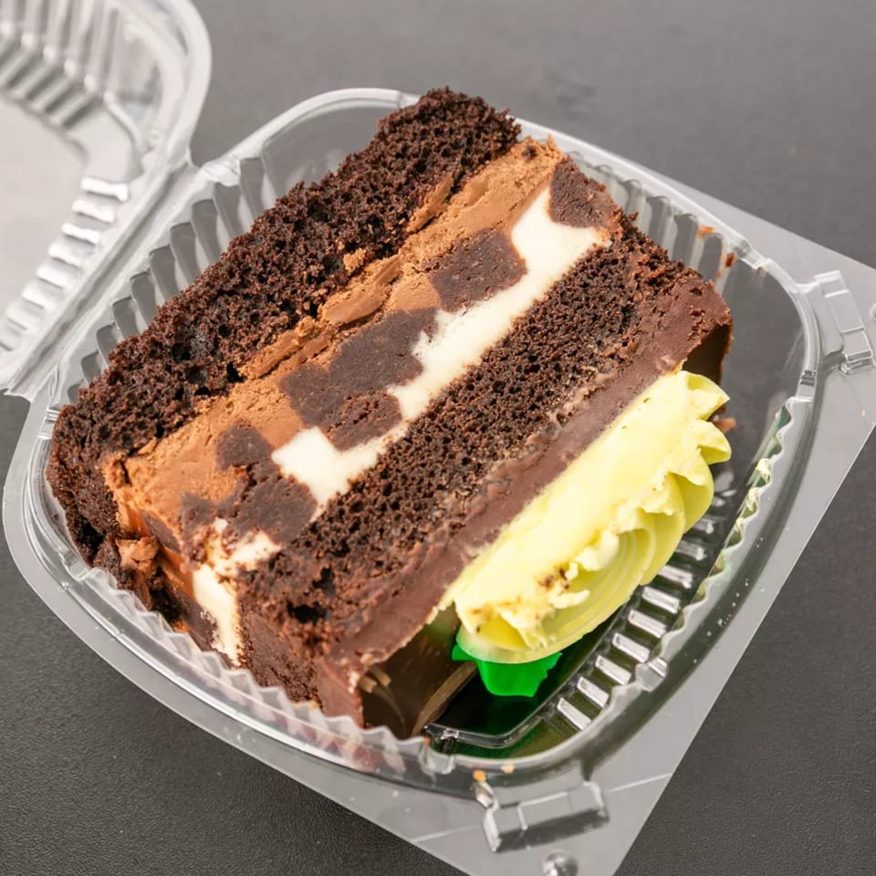 A slice of chocolate cake with cream cheese and chocolate frosting in a clear plastic container.
