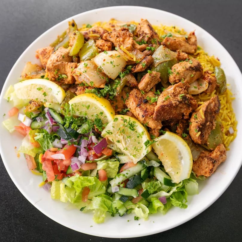A white plate with chicken and vegetable stir-fry over yellow rice, with a side salad and lemon slices.