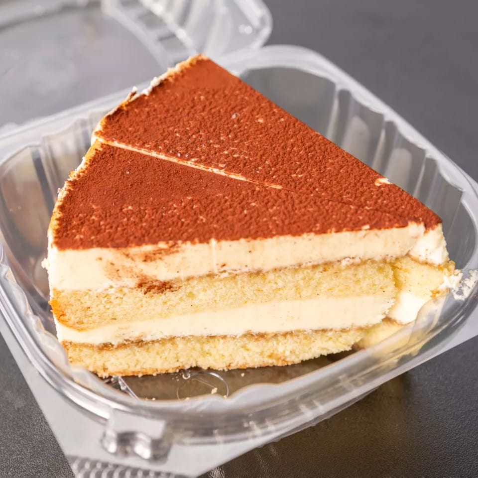 A slice of tiramisu with cocoa powder dusting, layered with cream and cake, in a plastic container.